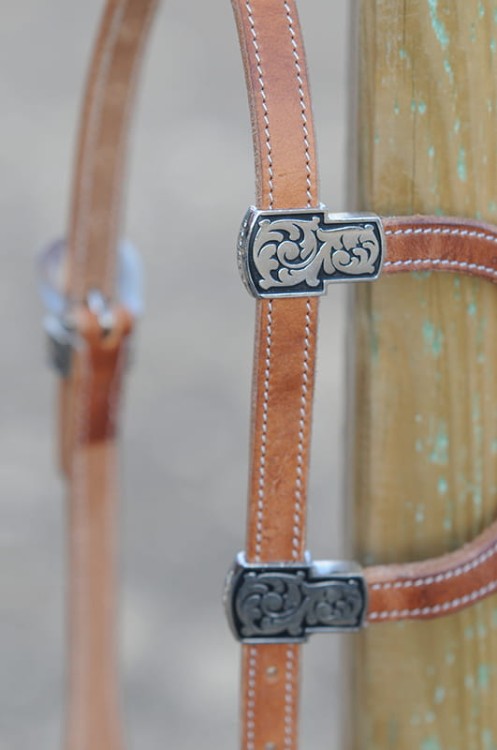 Detal prostokątnych keepers grawerowanych scrollwork na ogłowiu western — czarne tło, srebrna ramka, motyw roślinny